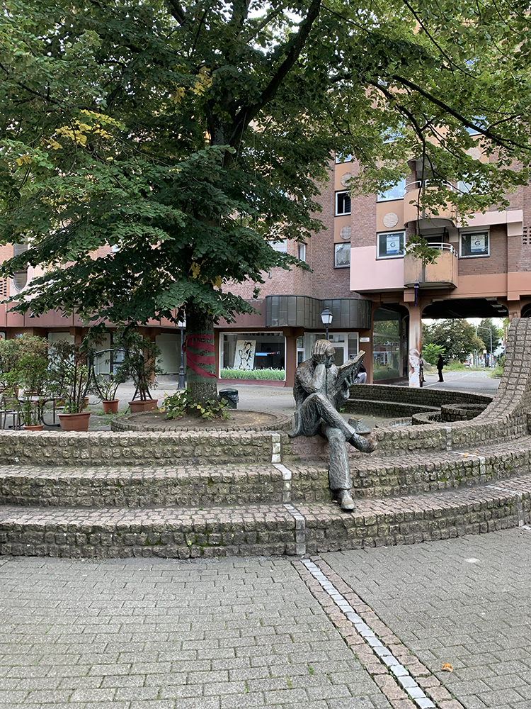 Bronzeskulptur Lesender, das Kunstwerk stellt einen sitzende Mann dar, der in der linken hbf ein Buchhhält in dem er liest, die rechte Hand ist zum Kopf geführt. Er sitzt auf gepflastertem Wall.
