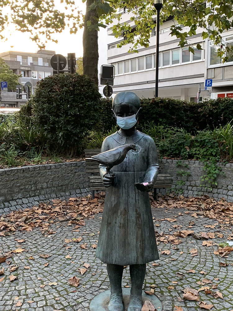 Bronzestatue Mädchen mit Taube in der rechten Hand. hier wurde eine chirugische Maske umgebunden ... im Hintergrund Sträucher und Gebäude