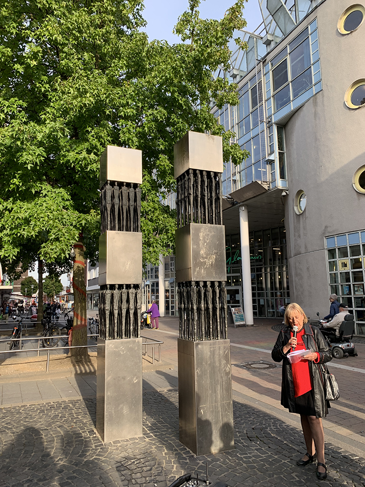 Im Vordergrund die Stehlen mit den Bronzeskulpturen des Monuments Miteinander. Die Stehlen sind aus Stahl, die Bronzeskulptutren sind männliche und weibliche Körper im Quadrat gesetzt, je 4 Stück pro Reihe, das Ganze je 2 Mal. Im Hintergrund ein Teilausschnitt des Glashaus Herten, in dem sich die Bibliothek befindet sowie die Rotunde für Veranstaltungen