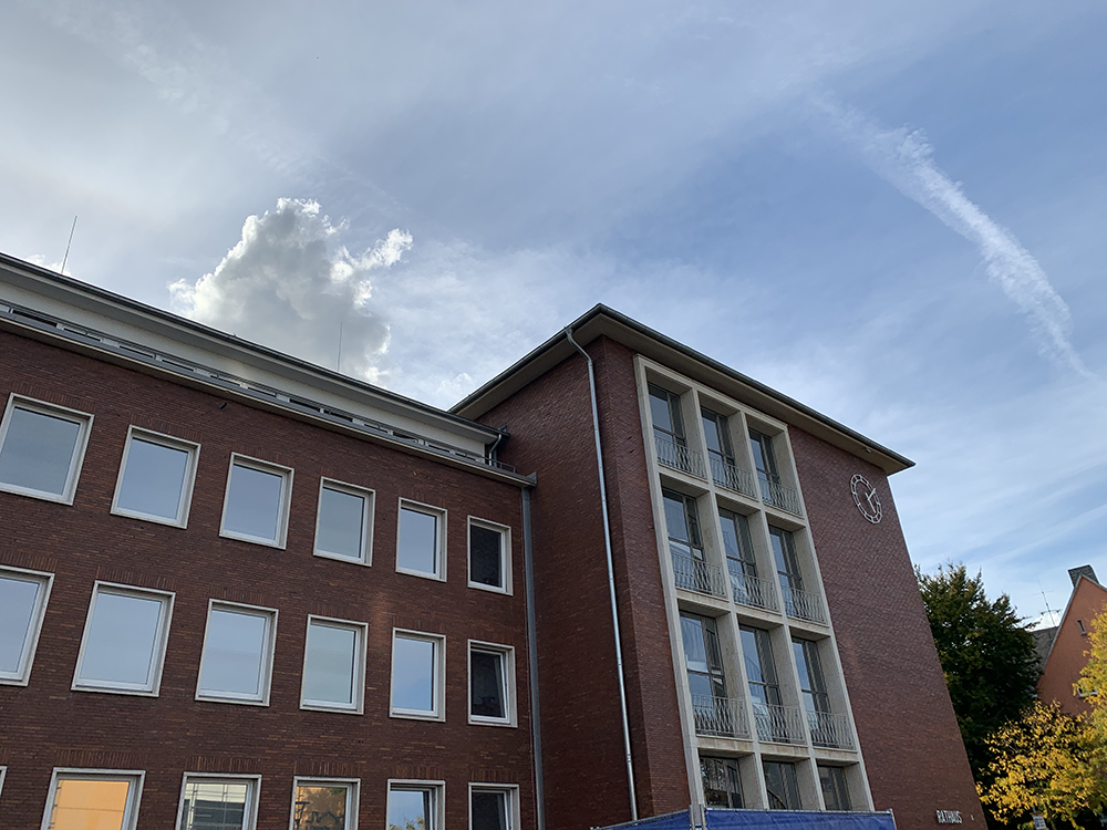 Block auf die Frontseite des Rathauses in Herten, brauner backsteinähnliche Fassade, Fenster in drei Reihen, rechts der vorgezogenen Eingangsbereich mit der Wendeltreppe. Die Uhr geht seit Jahren falsch. Blauer Himmel mit ein pass Wolken, man kann den Sonnenschein sehen.