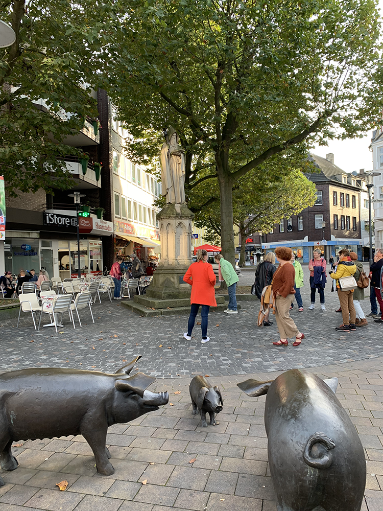 Eine Gruppe Teilnehmer:innen vor dem Denkmal Sankt Antonius, im Vordergrund eine kleine Schweineherde aus Bronze. Links eine Eisdiele mit Außenbestuhlung